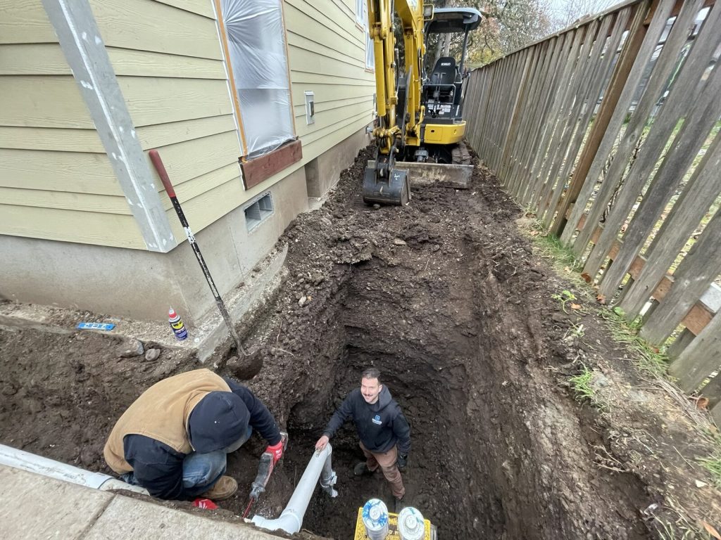 Open trench beside a house with a mini excavator and two Precision Plumbing technicians working in it