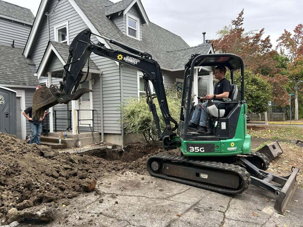 Precision Plumbing operator running a mini excavator beside a house during a sewer dig