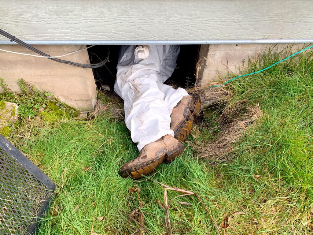Precision Plumbing technician in protective coveralls entering the crawl space beneath a home