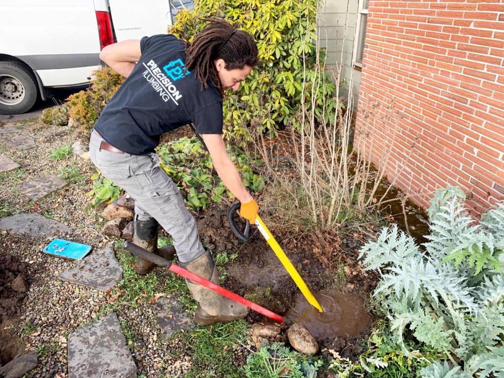 Precision Plumbing technician in a branded shirt digging out a leaking water line beside a brick home