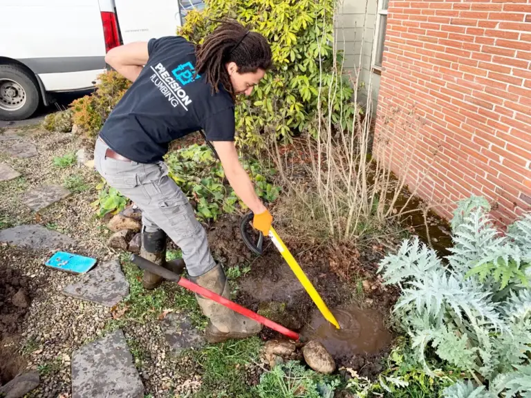 Precision Plumbing technician in a branded shirt digging out a leaking water line beside a brick home